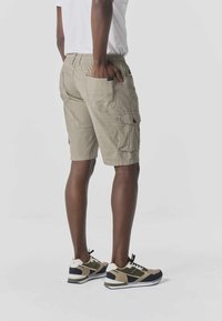 Moški, oblečen v bež cargo kratke hlače in večbarvne superge, stoji z eno roko v zadnjem žepu na sivi podlagi.