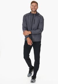 Graue Herrenjacke mit Kapuze und Reißverschluss, ausgestattet mit einer Brusttasche und elastischen Bündchen, kombiniert mit schwarzen Hosen und grauen Sportschuhen.
