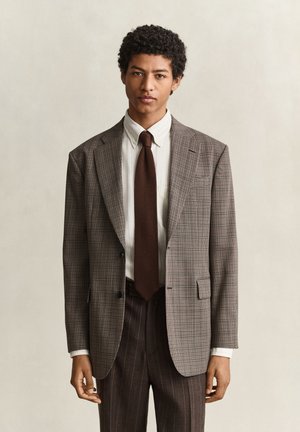 Junger Mann mit kurzen lockigen Haaren, der einen braunen karierten Blazer, ein weißes Hemd, eine dunkelbraune Krawatte und gestreifte dunkelbraune Hose trägt und vor einem neutralen Hintergrund steht.