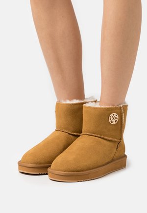 Bottes chevilles en daim beige avec une doublure intérieure en fourrure blanche douce et un accent de logo rond ; bout arrondi et semelle en caoutchouc plate avec texture antidérapante.