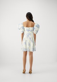 Vestido branco com padrão floral azul, design à mostra dos ombros, mangas bufantes e saia rodada. Par de sandálias de salto alto em tom de areia.