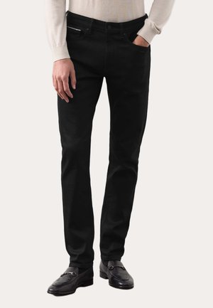 Mand iført sorte slim-fit bukser, beige langærmet skjorte og sorte loafers med metal detalje, står med den ene hånd i lommen.
