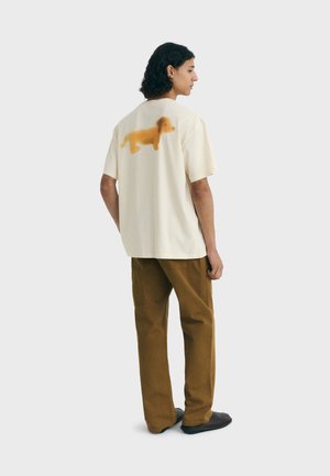 T-shirt crème à manches courtes avec une grande silhouette de chien orange au dos. Assorti à un pantalon marron, se tenant contre un fond clair.