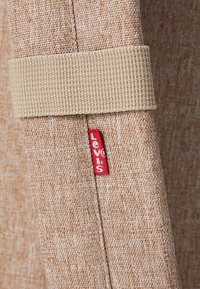 Rideau texturé beige avec un lien en tissu beige et une petite étiquette rouge de la marque Levi's cousue dans la couture.