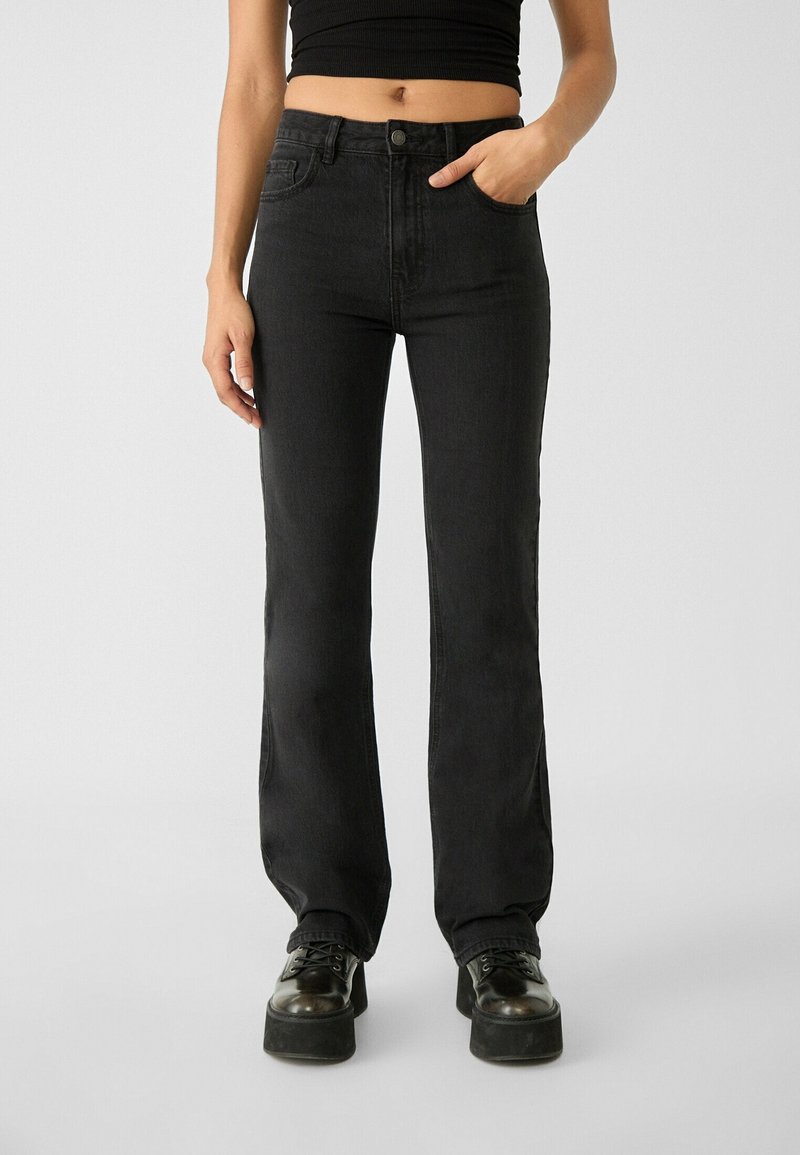 Femme portant un jean noir évasé taille haute, un top noir court et des chaussures noires à plateforme lacées, debout avec une main dans la poche.