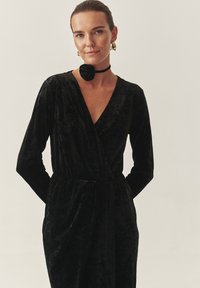 Črna žametna obleka z dolgimi rokavi, oblikovana v zavitju in s hudih pasom. Model nosi črn choker s podrobnostjo vrtnice in zlate uhane.