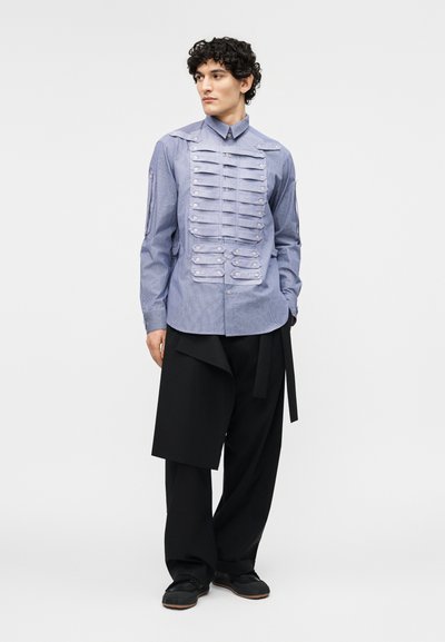 Homme portant une chemise bleue boutonnée avec des rabats plissés horizontaux et un pantalon large noir avec un panneau asymétrique, debout devant un fond uni.