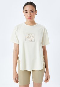 Lys creme t-shirt med korte ærmer, en rund kant, med et firkantet "NAMASTE" grafik i guld. Kombineret med beige cykelshorts.