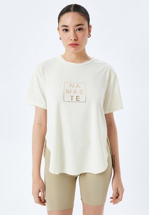 Lys creme t-shirt med korte ærmer, en rund kant, med et firkantet "NAMASTE" grafik i guld. Kombineret med beige cykelshorts.
