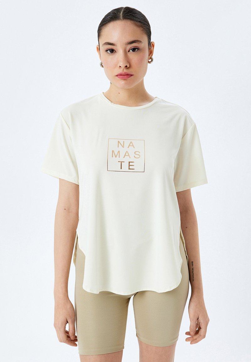 Lys creme t-shirt med korte ærmer, en rund kant, med et firkantet "NAMASTE" grafik i guld. Kombineret med beige cykelshorts.