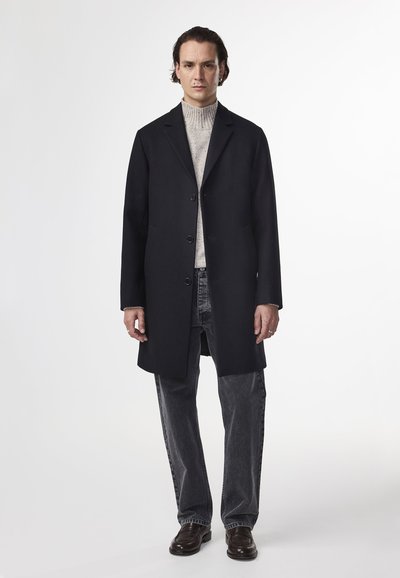 Musta villakangastakki, jossa on suora leikkaus, kolme nappia ja sivutaskut, pukeutuneena vaaleanharmaaseen ribbikauluspaitaan ja tummiin farkkuihin.