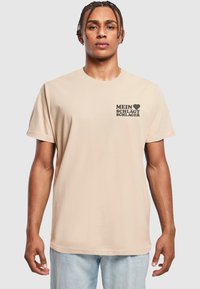 Beige bomulds-t-shirt med en sort grafiktryk, der står "MEIN ♥ SCHLÆGT SCHLAGER" på venstre brystområde. Korte ærmer og afslappet pasform.