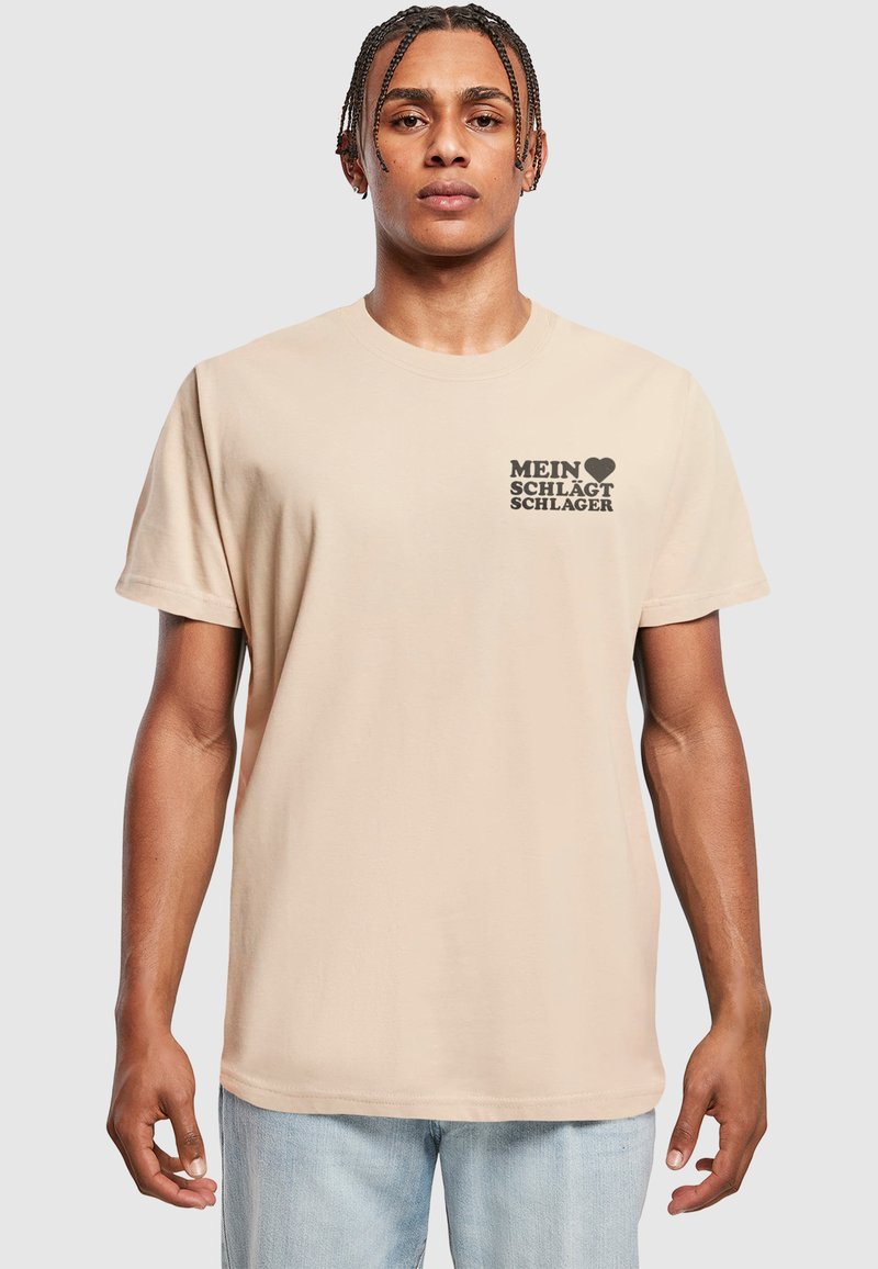 Beige bomulds-t-shirt med en sort grafiktryk, der står "MEIN ♥ SCHLÆGT SCHLAGER" på venstre brystområde. Korte ærmer og afslappet pasform.
