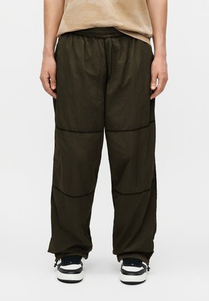 Person, die lockere dunkel olivgrüne Hosen mit sichtbaren Nähten und schwarz-weiße Sneakers trägt, steht vor einem einfarbigen Hintergrund.