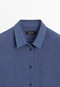 Blue long-sleeve linen shirt with pointed collar and black buttons, showing top section and brand label inside collar.