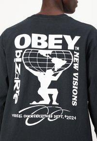 Schwarzes Langarmshirt mit weißem Grafikdruck. Das Design beinhaltet einen Globus, eine Figur, die ihn hebt, den Markennamen und Textelemente auf der Rückseite.