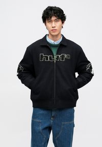 Casaco preto com fecho éclair e logótipo "huf" na frente, com detalhes contrastantes nas mangas. Usado sobre um suéter verde e uma camisa em camadas.