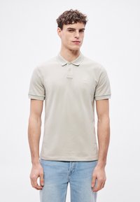 Ανοιχτό μπεζ πόλο πουκάμισο με γιακά, κοντά μανίκια και ένα μικρό λογότυπο στο στήθος, σε συνδυασμό με ανοιχτό μπλε τζιν. Ομαλή υφή.