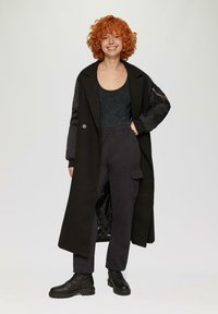 Langer schwarzer Mantel mit strukturiertem Design, kombiniert mit dunklen Cargo-Hosen und schwarzen Schnürstiefeln. Texturiertes Innenfutter ist sichtbar.