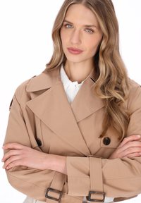Beige trenchcoat med breda slag, knapplist och ärmslut med skärp. Slät tygstruktur, bärs över en vit skjorta med krage.