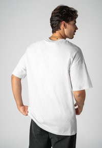 Weißes Baumwoll-T-Shirt, lockere Passform, kurze Ärmel, Rundhalsausschnitt, glatte Textur, getragen zu dunklen Jeanshosen, von hinten dargestellt.