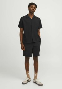 Chemise noire à manches courtes et shorts assortis en tissu texturé. La personne porte des baskets beiges avec des rayures noires et des chaussettes marron.