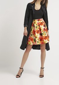 Abrigo largo negro sobre una blusa sin mangas negra, combinado con una falda plisada multicolor con estampado floral y sandalias negras de tacón alto.