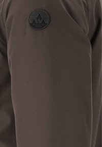 Braunes Stoffpatch mit einem runden schwarzen Logo, das Berge und Textdetails zeigt, an der Seite eines Kleidungsstücks angenäht. Glatte Textur.