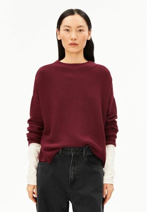 Femme aux longs cheveux noirs porte un pull ample bordeaux sur une chemise blanche à manches longues et un jean noir taille haute sur un fond uni.