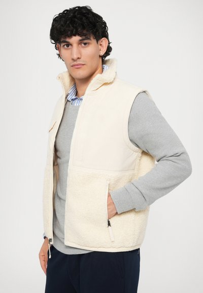 Bežová fleece vesta s hladkým vrškem, zapínáním na zip, bočními kapsami a měkkou texturou. Nosená přes světle šedý svetr.