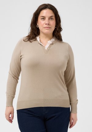 Kvinde med mørkt krøllet hår iført beige langærmet sweater over hvid skjorte med blonderkrave og mørkeblå bukser på en ensfarvet baggrund.