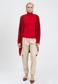 Rød turtleneck sweater med en fuzzy tekstur, båret under en lang rød cardigan. Beige cargo bukser med sidelommer og ankler. Brune flade sko.