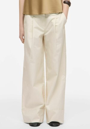 Personne portant un pantalon crème large et ample avec des plis devant, des chaussures noires, un haut marron et un bracelet marron foncé à un poignet.