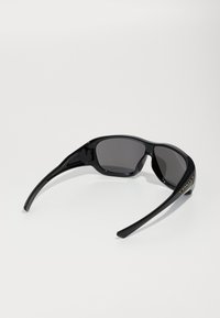 Black sunglasses with a sleek design, gray lenses, and a noticeable logo on the side. The frame features textured accents for grip.