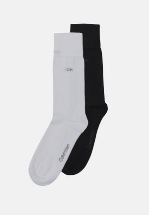 Deux paires de chaussettes, une blanche et une noire, au design minimaliste. Chaque chaussette arbore un petit logo "CK" en couleur contrastante.