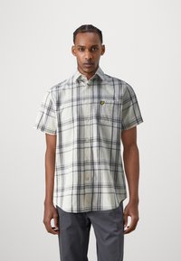 Camisa de cuadros de manga corta en beige con rayas grises y marinas, que presenta un cierre frontal con botones y un bolsillo en el pecho izquierdo con un logo amarillo.