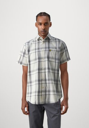 Camisa de cuadros de manga corta en beige con rayas grises y marinas, que presenta un cierre frontal con botones y un bolsillo en el pecho izquierdo con un logo amarillo.