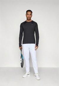 Zwarte atletische lange mouwen shirt met accentstiksels, gecombineerd met witte broeken en witte sneakers. Houdt een tennisracket vast.