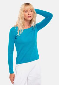 Türkises Longsleeve-Oberteil aus weichem Strickmaterial, mit Rundhalsausschnitt und taillierten Ärmeln, kombiniert mit weißen, weit geschnittenen Hosen.