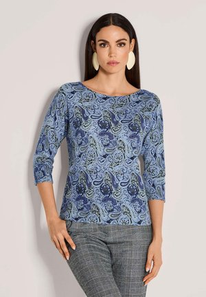 Vrouw met lang donker haar die grote crèmekleurige oorbellen draagt, een blauw paisley-patroon bovenstuk met driekwartmouwen en een grijs geruit broek.