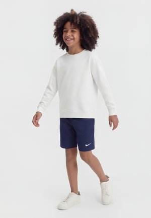 Usmievajúce sa dieťa s kučeravými vlasmi kráča, má na sebe bielu mikinu, tmavomodré šortky značky Nike a biele tenisky na jednoduchom bielom pozadí.