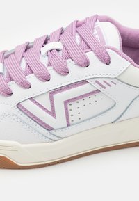 Fehér sneaker, lila fűzőkkel, textúrált bőr felsőrésszel, kiemelt logóval és perforált részletekkel az oldalain.