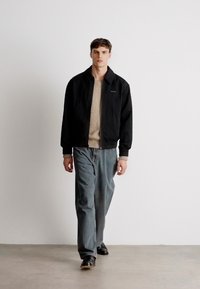 Ung mand, der går indendørs iført en sort jakke, beige sweater, blå jeans og sorte sko foran en simpel hvid baggrund.