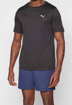 Homme portant un t-shirt de sport noir à manches courtes Puma et un short bleu, debout les bras détendus devant un fond uni.