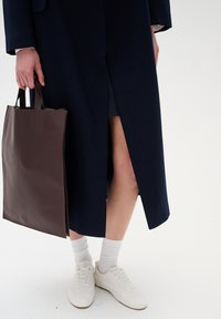 Cappotto di colore navy con uno spacco frontale, abbinato a una borsa tote in pelle marrone e sneakers bianche. L'outfit presenta tessuti testurizzati e colori a contrasto.