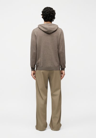 Άνδρας με σγουρά μαύρα μαλλιά, στέκεται με την πλάτη γυρισμένη, φορώντας καφέ φούτερ με κουκούλα και φαρδιά μπεζ παντελόνια, σε απλό φόντο.