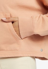 Sudadera melocotón con puño acanalado y un bolsillo frontal, que presenta detalles de costura sutiles y un pequeño acento de hardware plateado en forma de círculo.