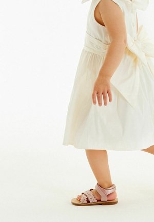 Robe beige clair avec un nœud à la taille ; sandales roses avec des accents floraux et une bride autour de la cheville. Tissu lisse et textures douces.