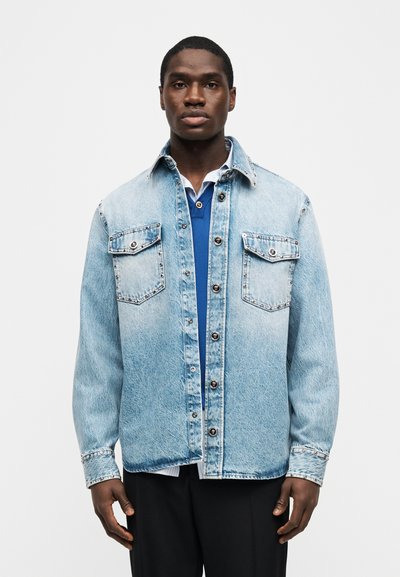 Άνδρας που κοιτάζει μπροστά, φοράει ένα ανοιχτό γαλάζιο τζιν μπουφάν με τρουκς πάνω από ένα στρωμένο μπλε και λευκό πουκάμισο, στέκεται μπροστά σε λευκό απλό φόντο.