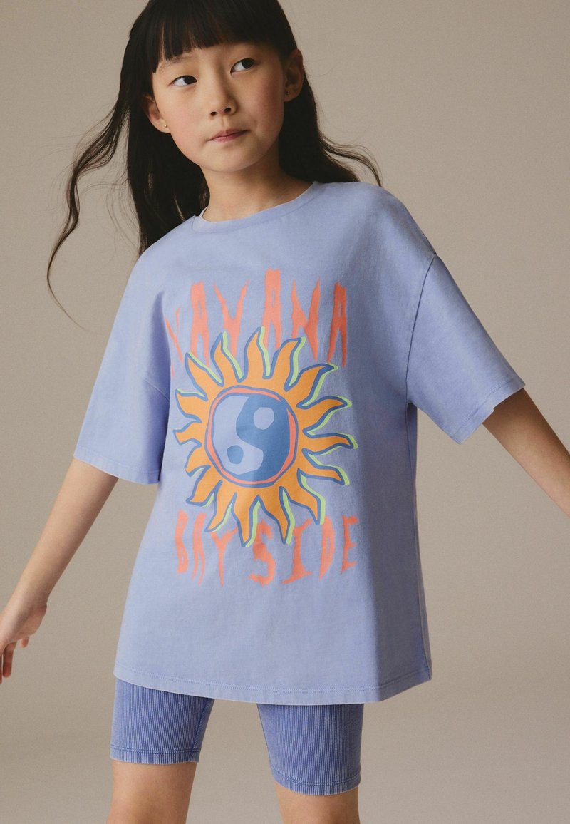 Giovane ragazza che indossa una maglietta oversize viola chiaro con un disegno di sole e yin-yang e pantaloncini da ciclismo a coste abbinati.